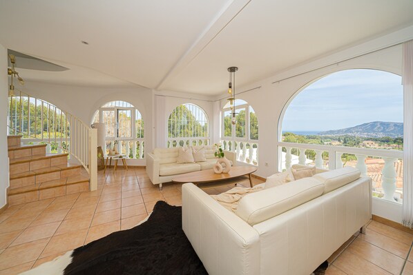 A spacious living room with a white sofa, a coffee table, and a black rug. There are large arched windows offering a view of the outdoors.