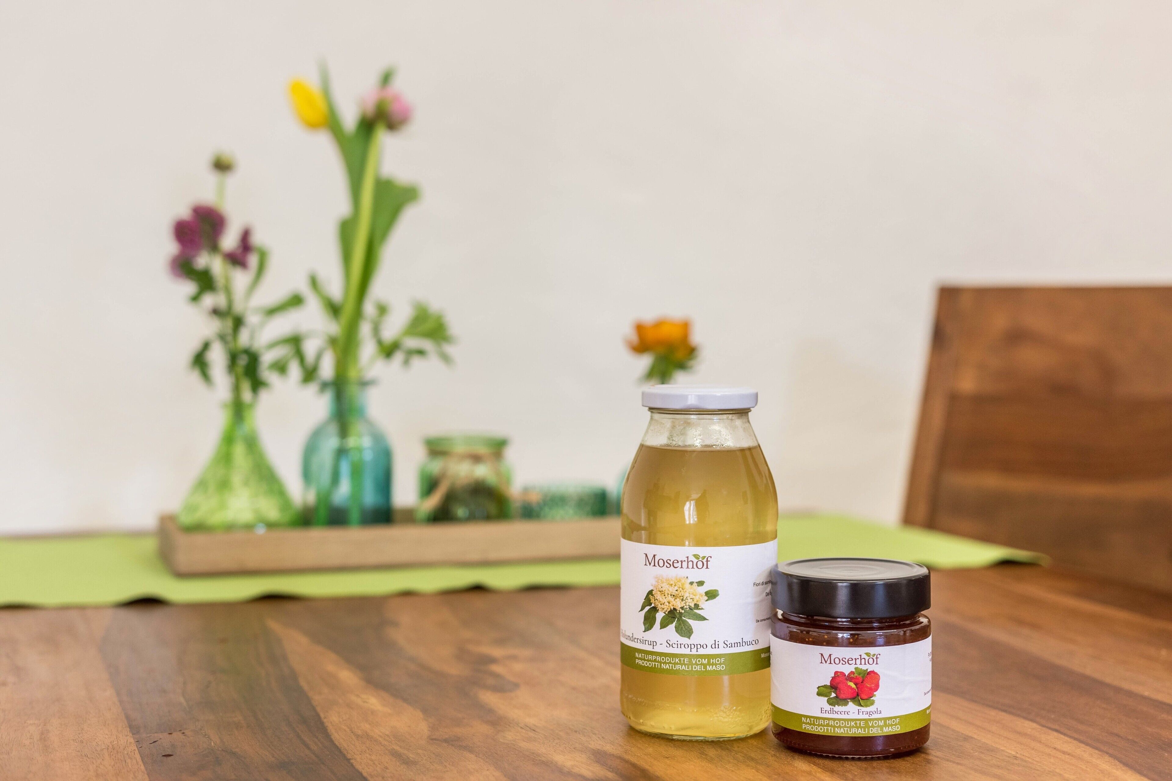 Une table en bois sur laquelle se trouvent une bouteille et un pot de produit Moserhof, un vase avec des fleurs et, au fond, une boîte en bois.