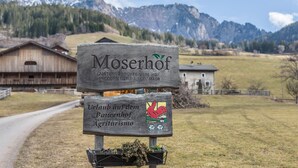 A wooden sign with text and a coat of arms, set against a rural landscape with mountains and buildings. - Studio 'Moserhof - Sunnseit' with Mountain View, Garden & Wi-Fi (Sarnthein)