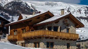 Un edificio in stile chalet con un balcone in legno, pareti in pietra e montagne sullo sfondo.