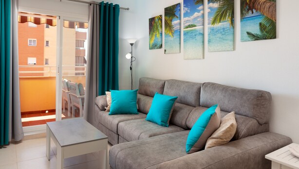 Ein Wohnzimmer mit grauem Sofa, türkisfarbenen Kissen, einem kleinen Tisch und einem Balkon mit Sitzgelegenheiten.