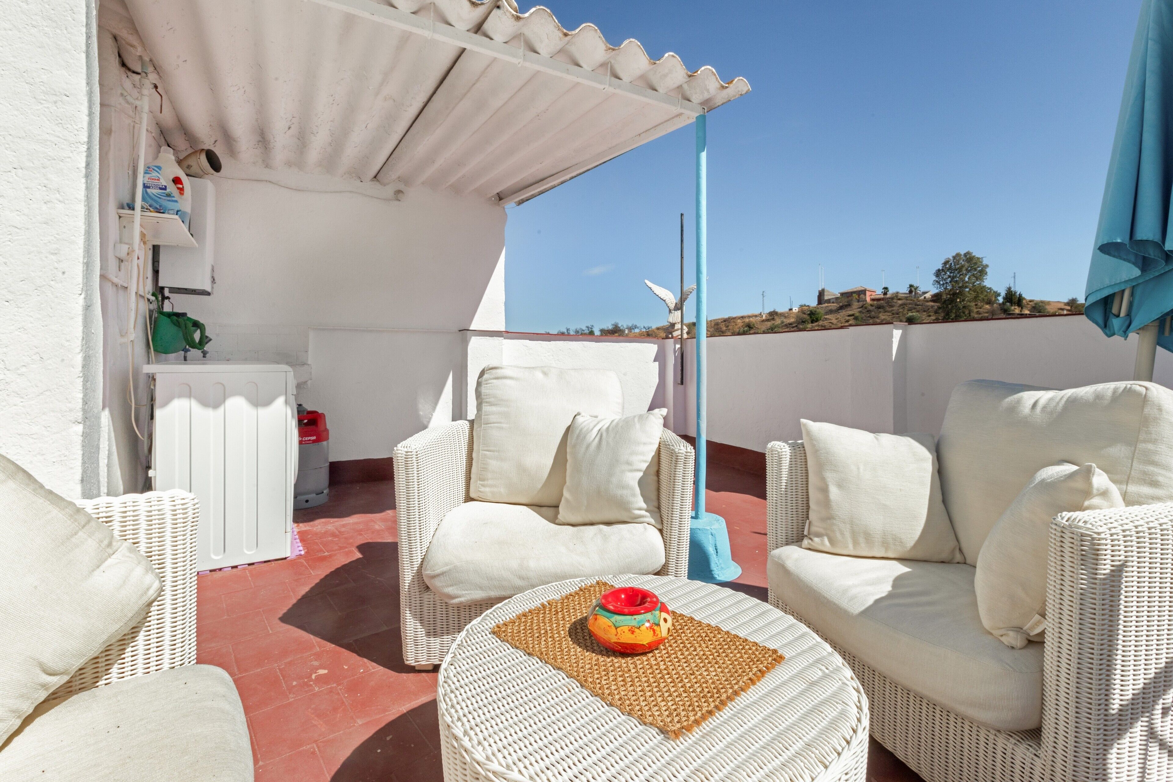 Una terraza en la azotea con mobiliario de mimbre blanco, una pequeña mesa con un objeto decorativo y vistas al paisaje.