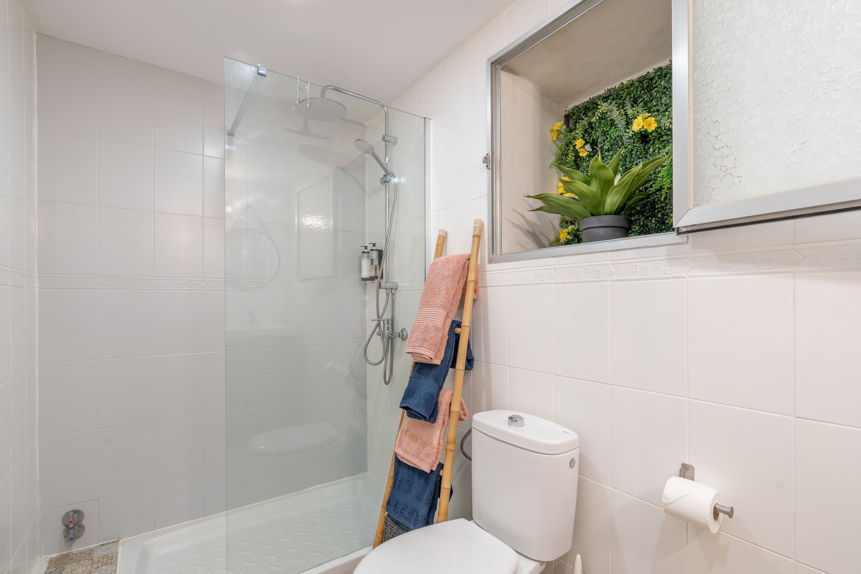 Un baño con cabina de ducha de vidrio, inodoro y una pequeña ventana con una planta.