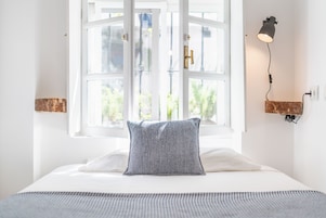 A neatly made bed with a patterned pillow, a window with white frames, and a wall-mounted lamp.