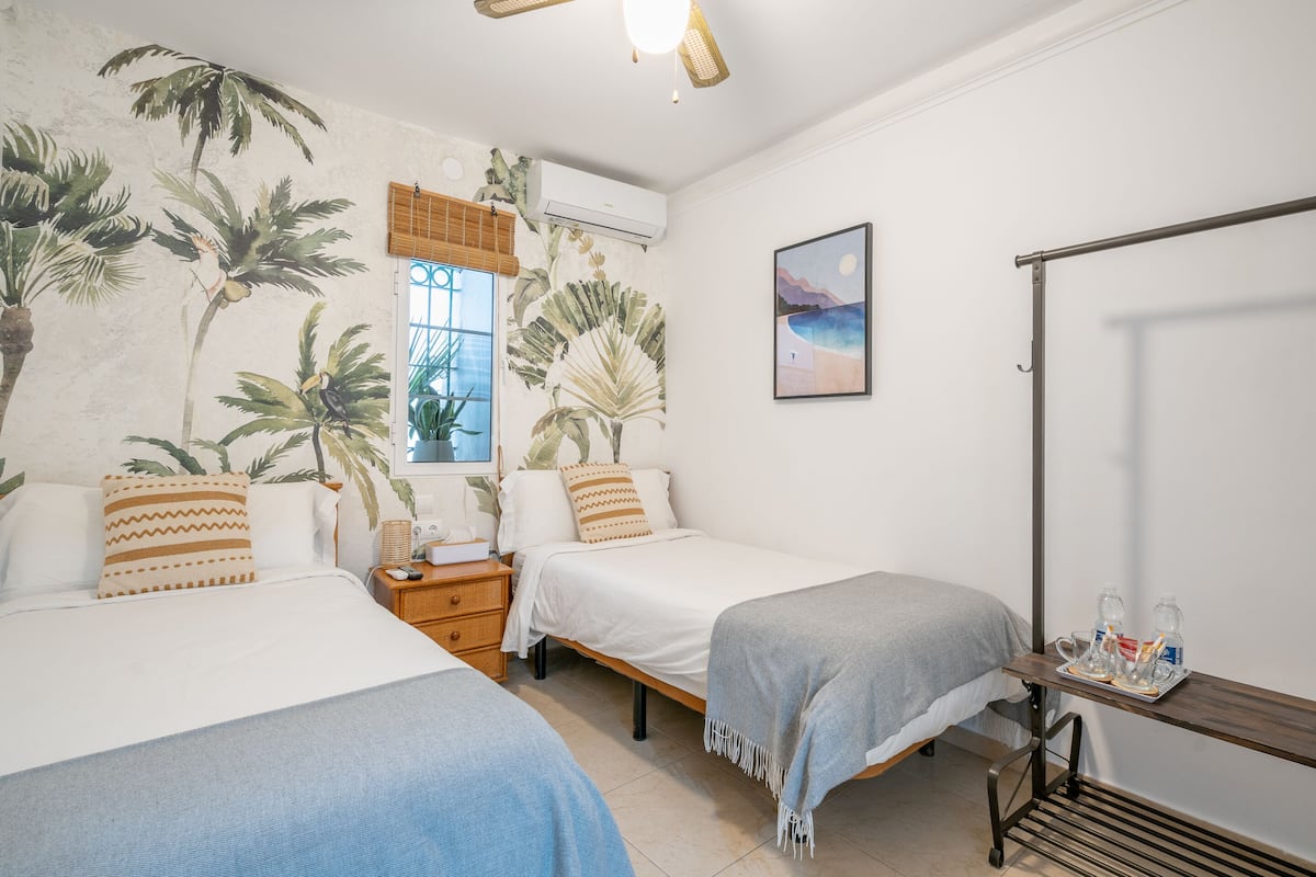 A room with two beds, a window with blinds, a wall-mounted air conditioner, a painting, and a small table with items.
