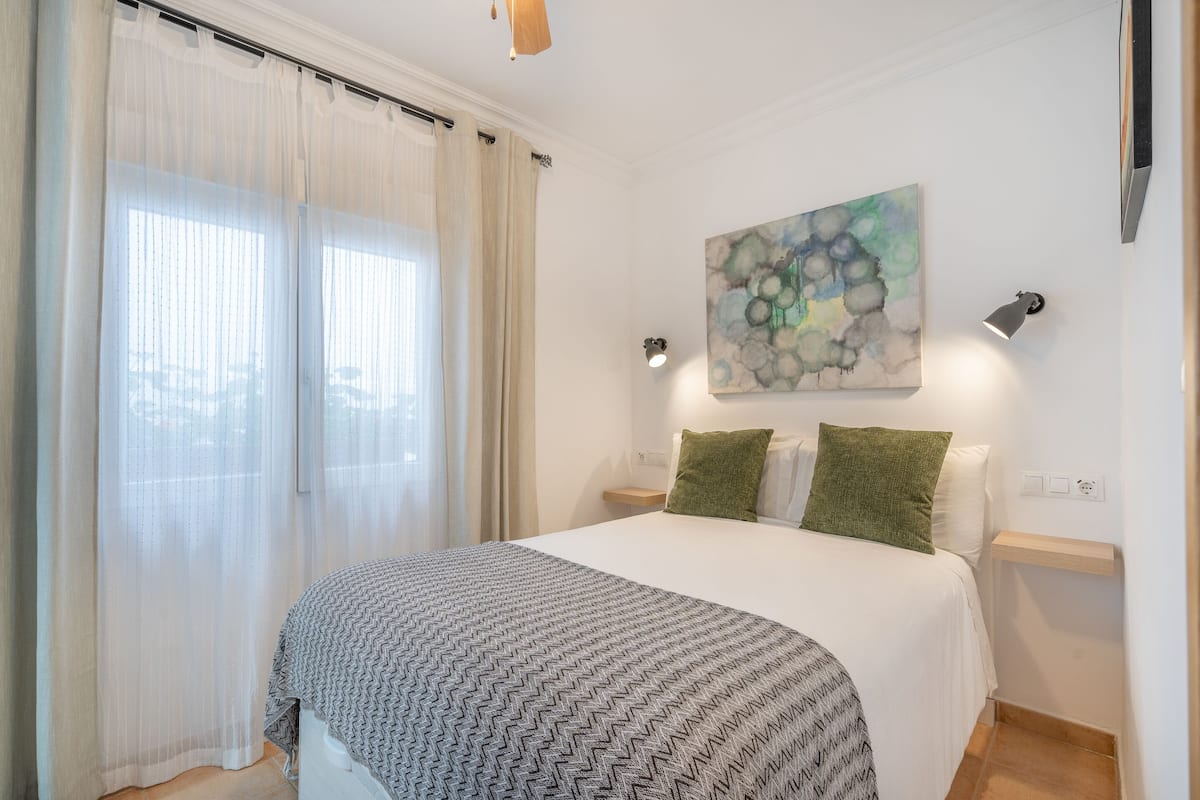 A bedroom with a bed, curtains, a wall art piece, and bedside lamps.