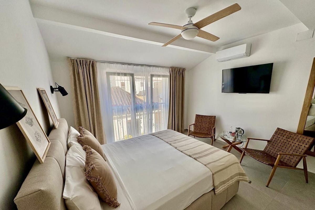 A hotel room with a bed, two chairs, a TV, a ceiling fan, and a window with curtains.