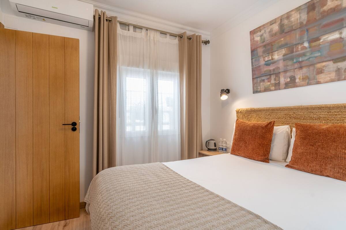 A bedroom with a bed, a wooden wardrobe, a nightstand with a kettle, and a painting on the wall.