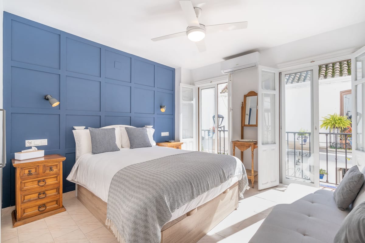A bedroom with a large bed, a dresser, a ceiling fan, and a balcony with a view.
