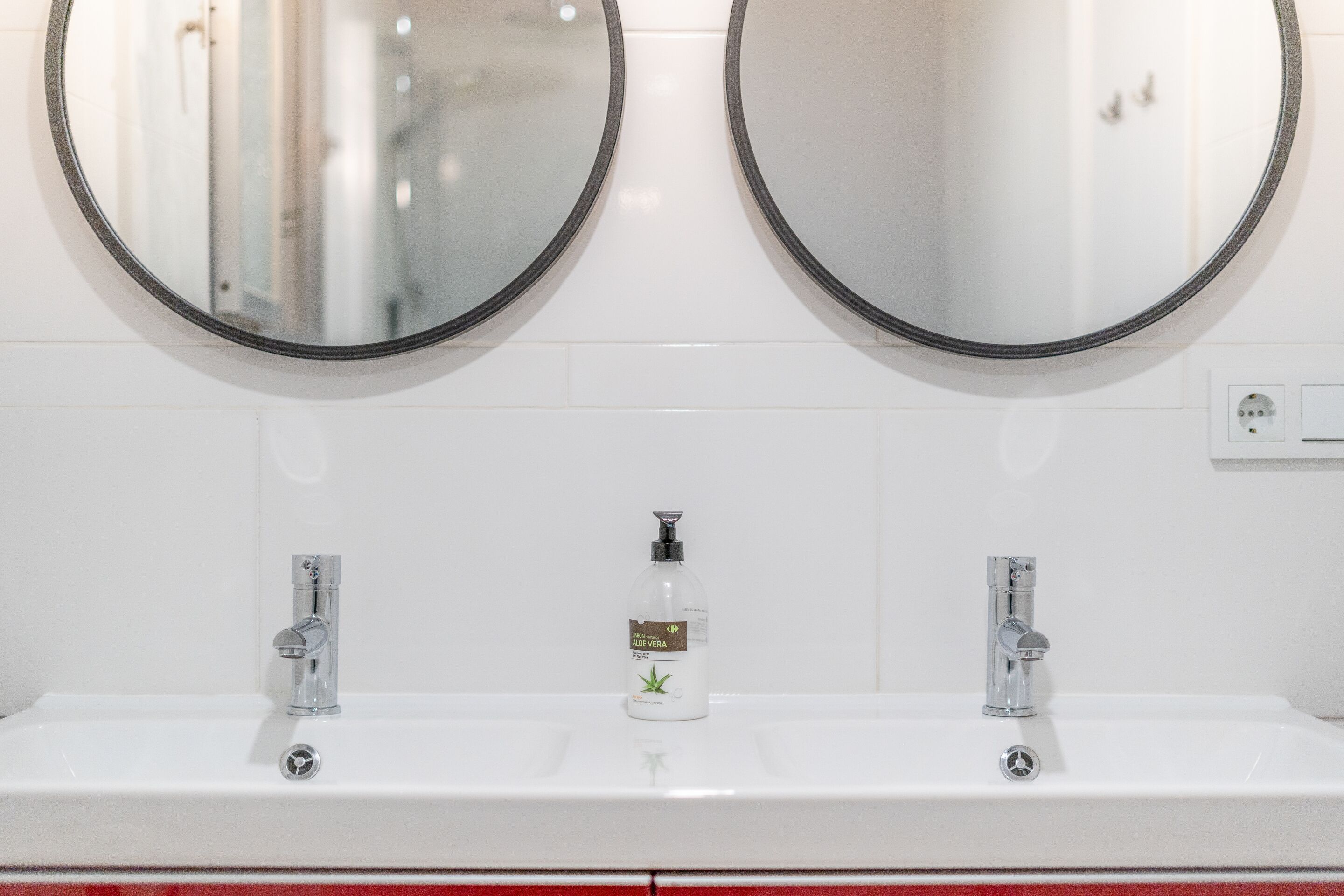Une salle de bain avec deux miroirs ronds, un lavabo blanc doté de deux robinets et un flacon de lotion à base d’aloe vera.