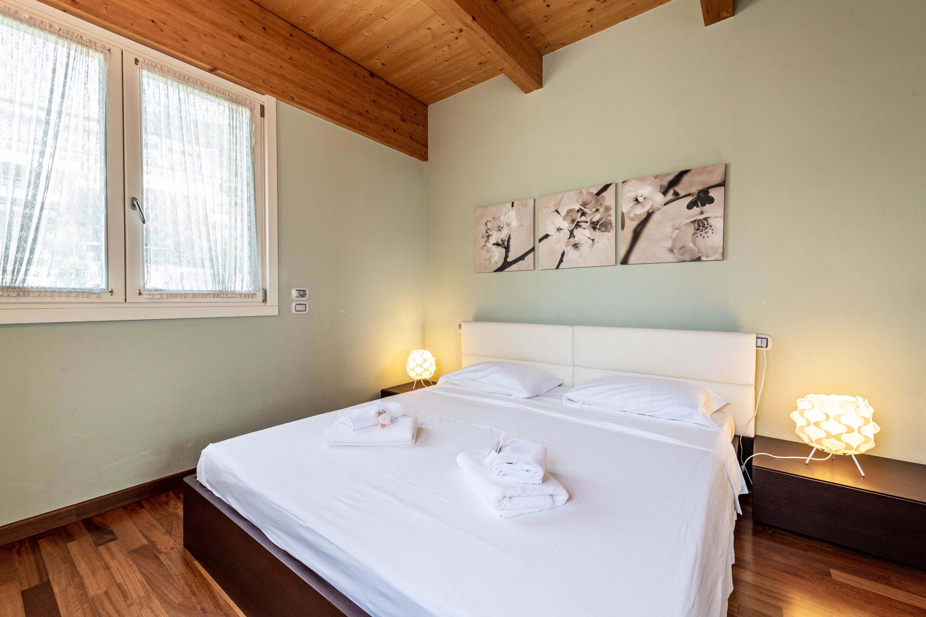 A neatly made bed with towels, wooden ceiling beams, a window with curtains, and wall art.