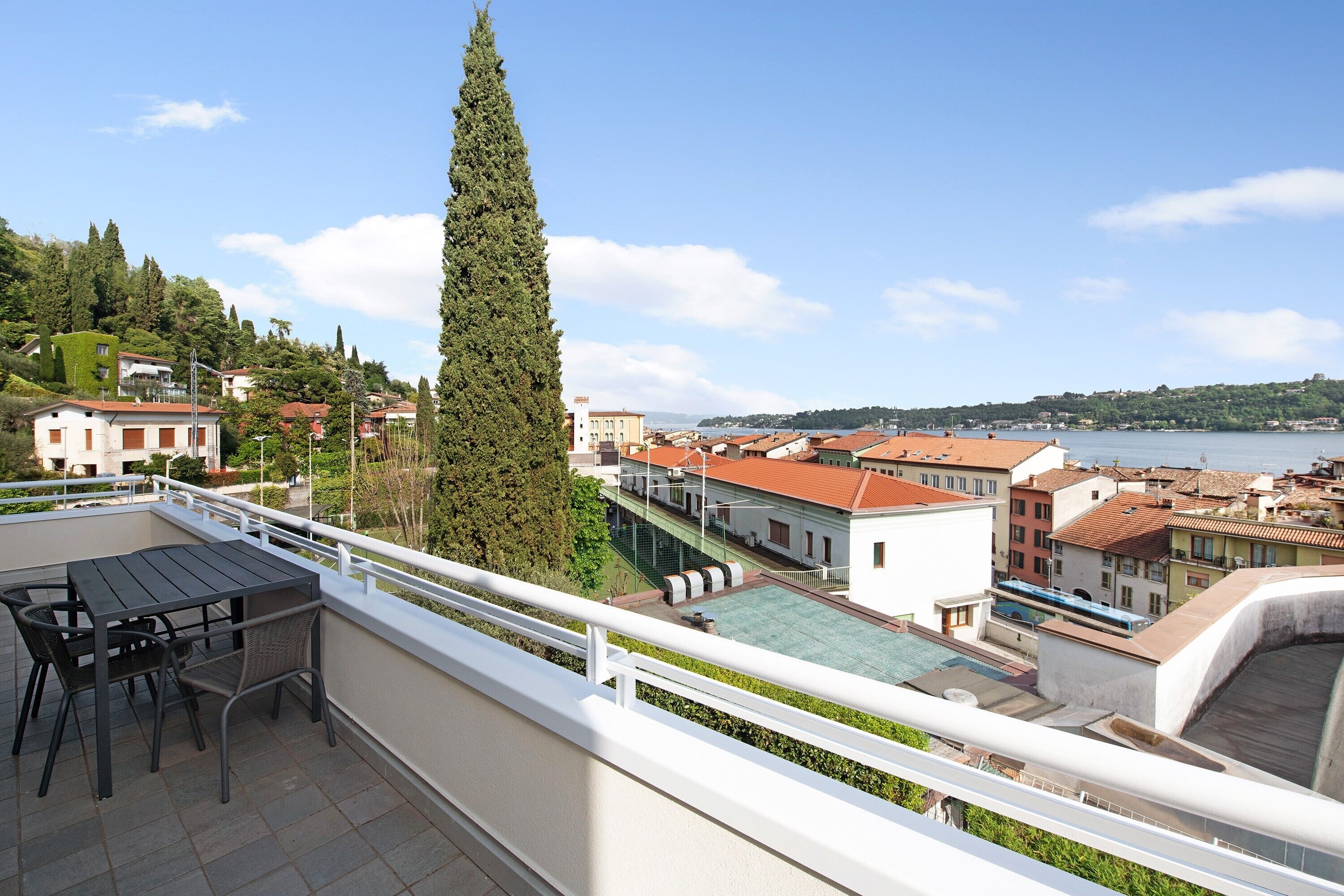 Un balcone con tavolo e sedie, affaccia su una cittadina con edifici e un corpo d'acqua.