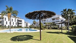 Un área de piscina con una palmera y un paraguas de paja, rodeada de césped cuidado y edificios modernos.