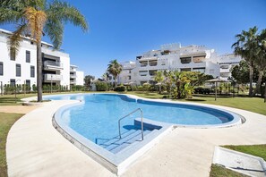 Une piscine avec un escalier en métal, entourée d’une terrasse en béton, avec des bâtiments blancs et des palmiers en arrière-plan.