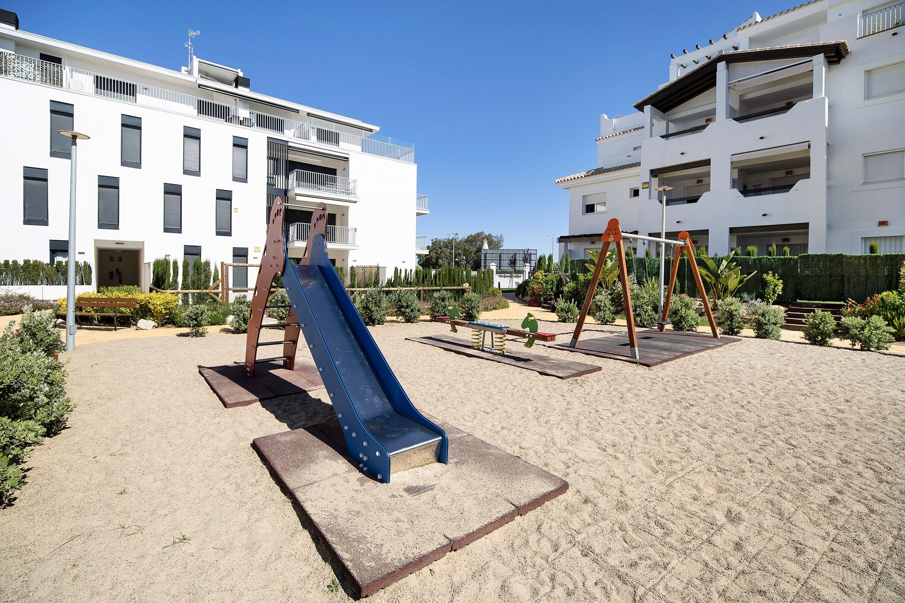 Ein Spielplatz mit Rutsche und Schaukeln, umgeben von Sand, mit Wohngebäuden im Hintergrund.