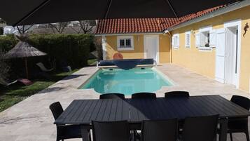 Uma área de piscina ao ar livre com mesa e cadeiras, um alpendre coberto e uma casa ao fundo.