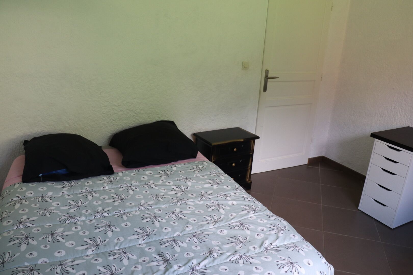 Ein Schlafzimmer mit einem Bett, zwei Kissen, einem Nachttisch, einer Kommode und einer Tür.