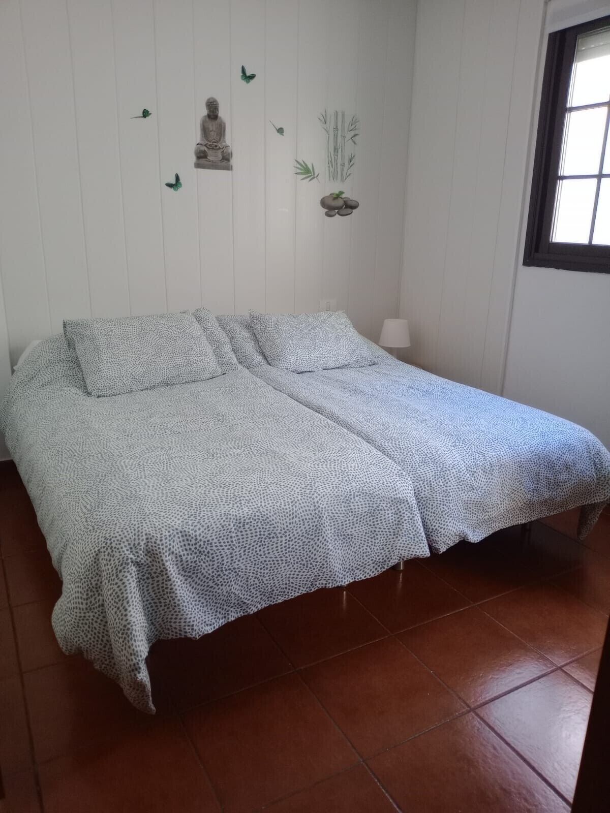 Un lit simple avec une couvre-lit à motifs, une statue de Bouddha fixée au mur et une petite plante.