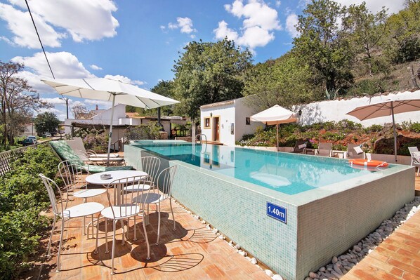 Una piscina con sillones de descanso y sombrillas, rodeada de follaje y una terraza con baldosas.