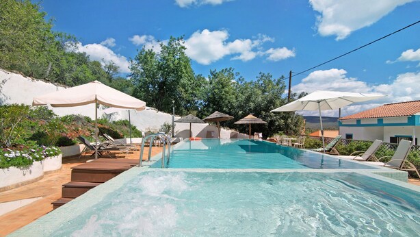A swimming pool with lounge chairs and umbrellas, surrounded by greenery and a clear sky.
