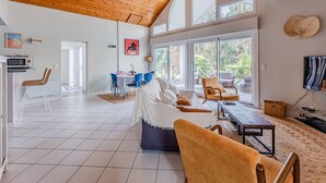 A spacious living area with a high wooden ceiling, a dining area with blue chairs, and large windows offering views of the outdoors. - Biscarrosse: Villa avec Piscine Privée, Jardin, 3 Chambres, Proche du Golf, WiFi Inclus (Biscarrosse)