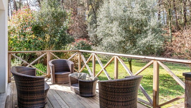 A wooden deck with wicker furniture and a railing overlooking a garden with trees and shrubs. - Biscarrosse: Villa avec Piscine Privée, Jardin, 3 Chambres, Proche du Golf, WiFi Inclus (Biscarrosse)