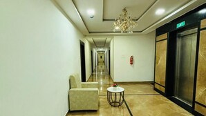 Un hall d’hôtel avec un sol en marbre, un canapé, une table ronde ornée d’un vase, un lustre, un ascenseur et un extincteur.