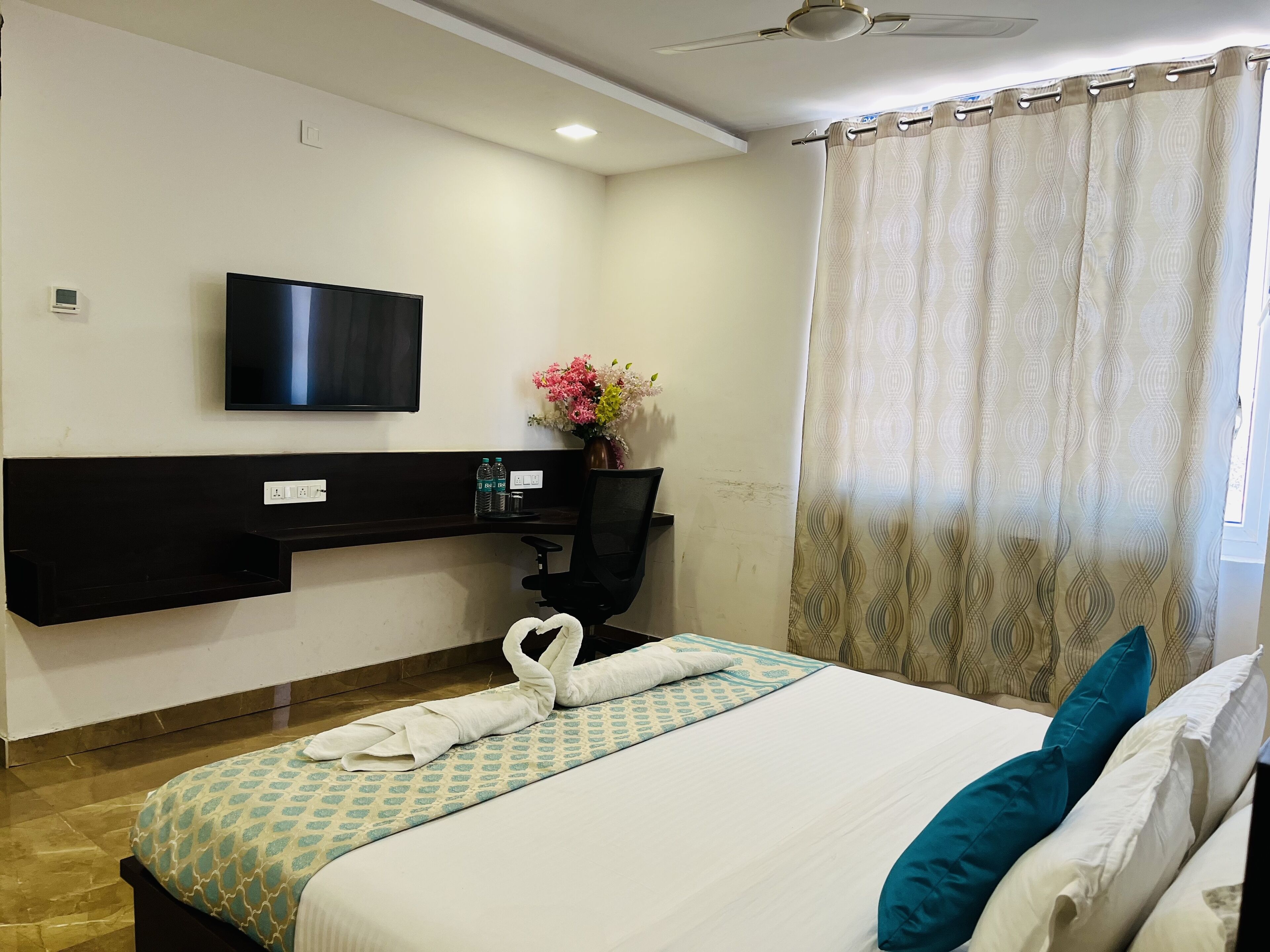 A hotel room with a bed, a flat-screen TV mounted on the wall, a desk with a computer, and a window with curtains.