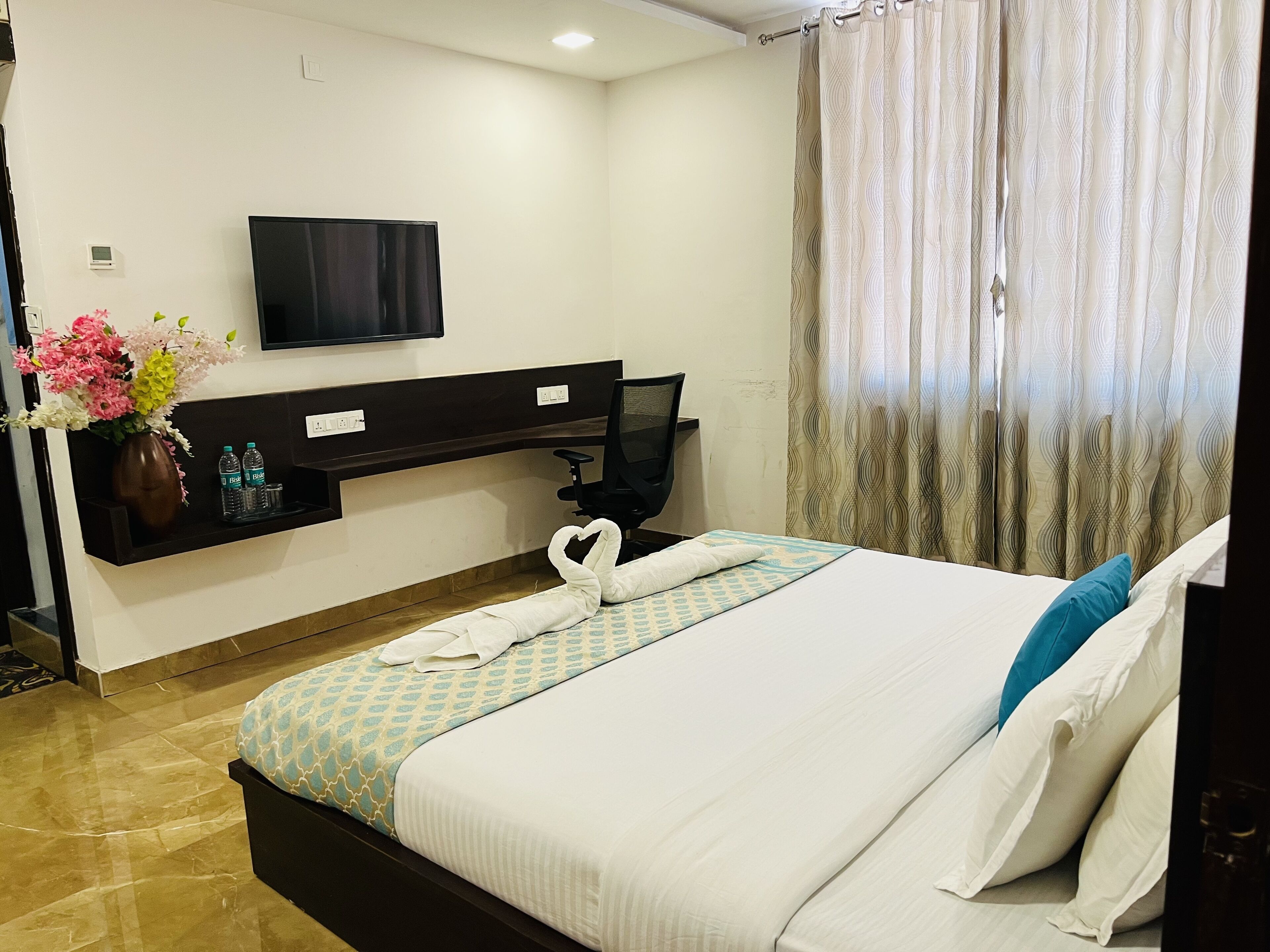 A hotel room with a bed, a desk with a computer, a flat-screen TV, a vase with flowers, and a window with curtains.