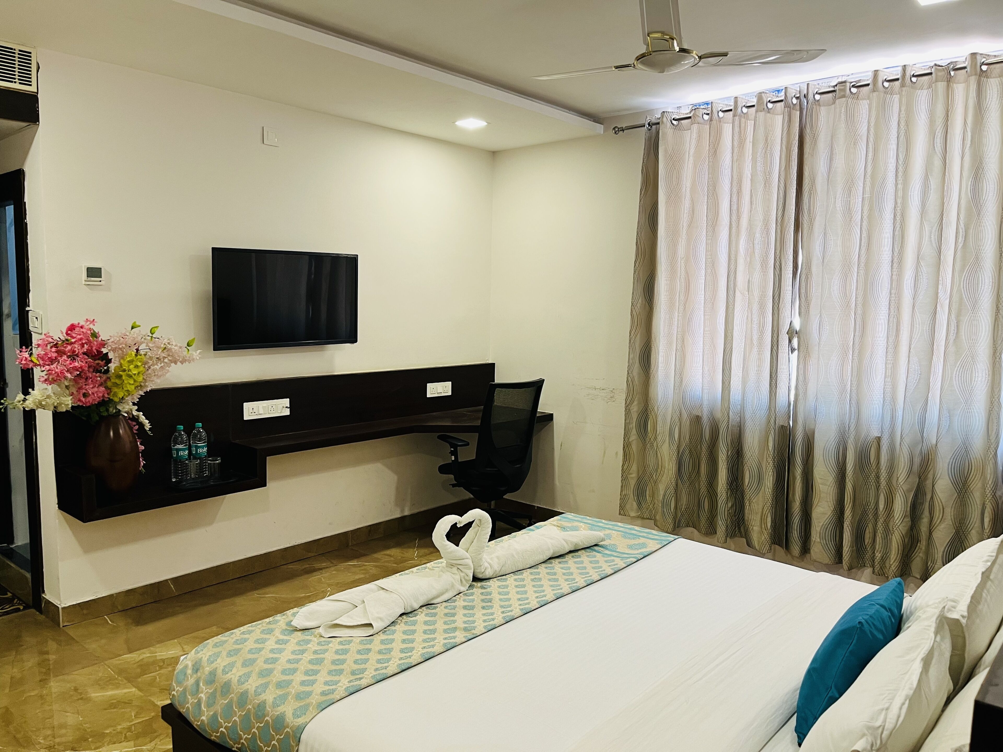 A hotel room with a bed, a desk with a computer, a flat-screen TV, a vase with flowers, and a window with curtains.