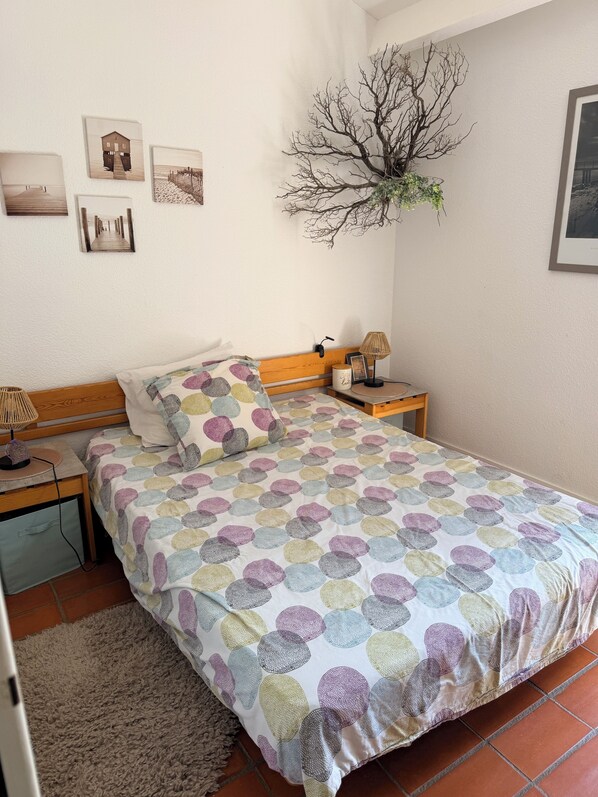A bedroom with a bed, a bedside table with a lamp, a wall-mounted plant, and framed pictures. - Cozy little house by the lake (Fitou)