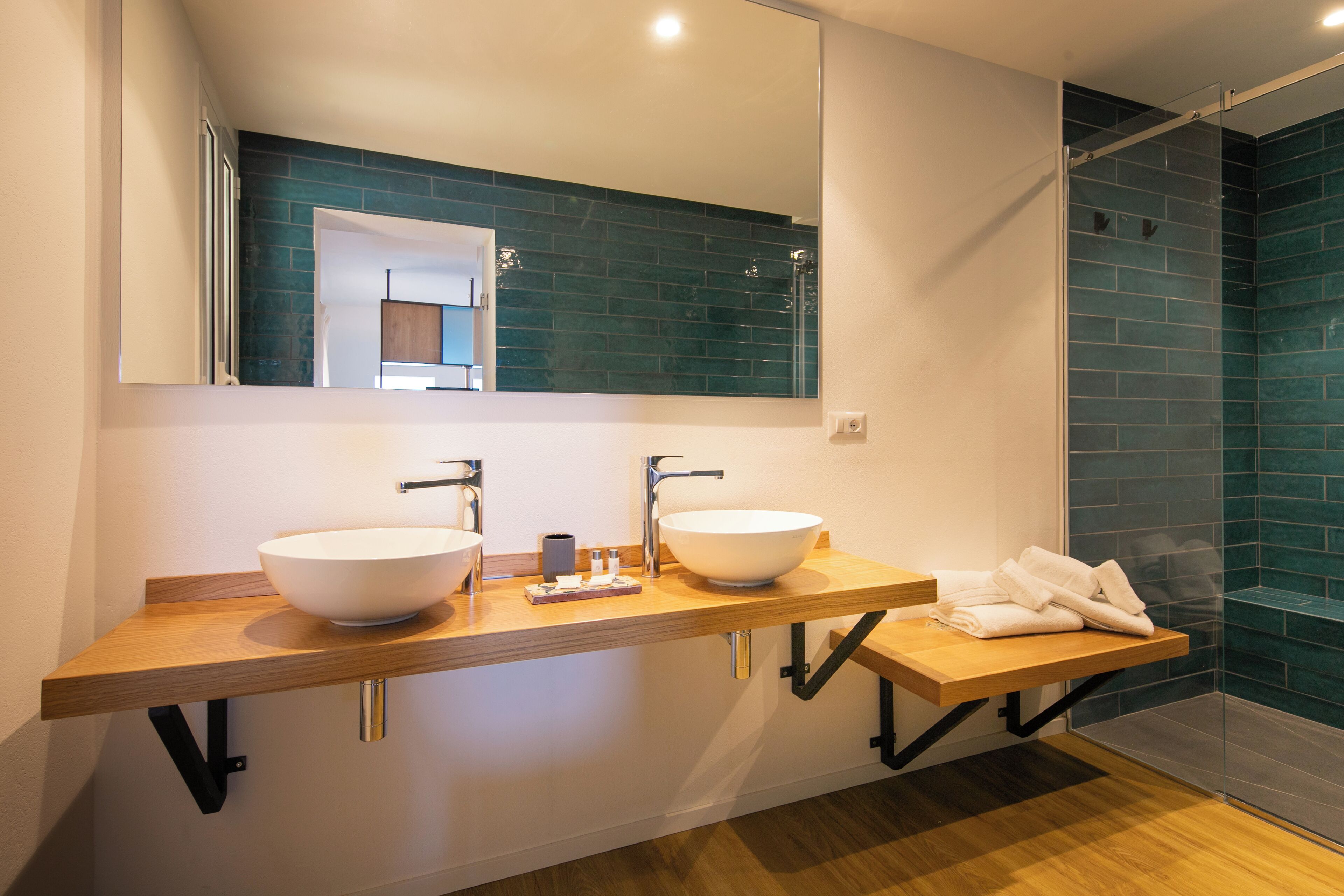 Un baño moderno con dos lavamanos grandes de color blanco, un mueble de tocador de madera, un espejo y una zona de ducha con azulejos azules.