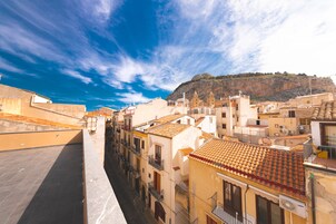 A narrow street lined with buildings featuring terracotta roofs and balconies, leading to a hill with a church.