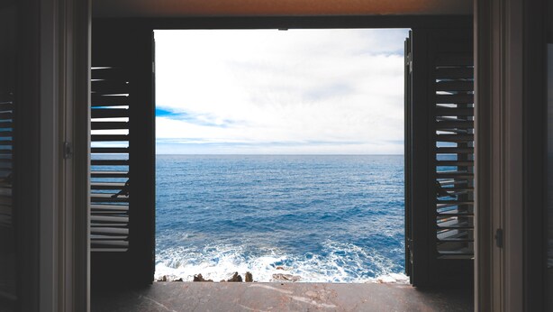 A view through a window with shutters, overlooking a body of water and rocky shoreline. - Taliammari (Cefalù)