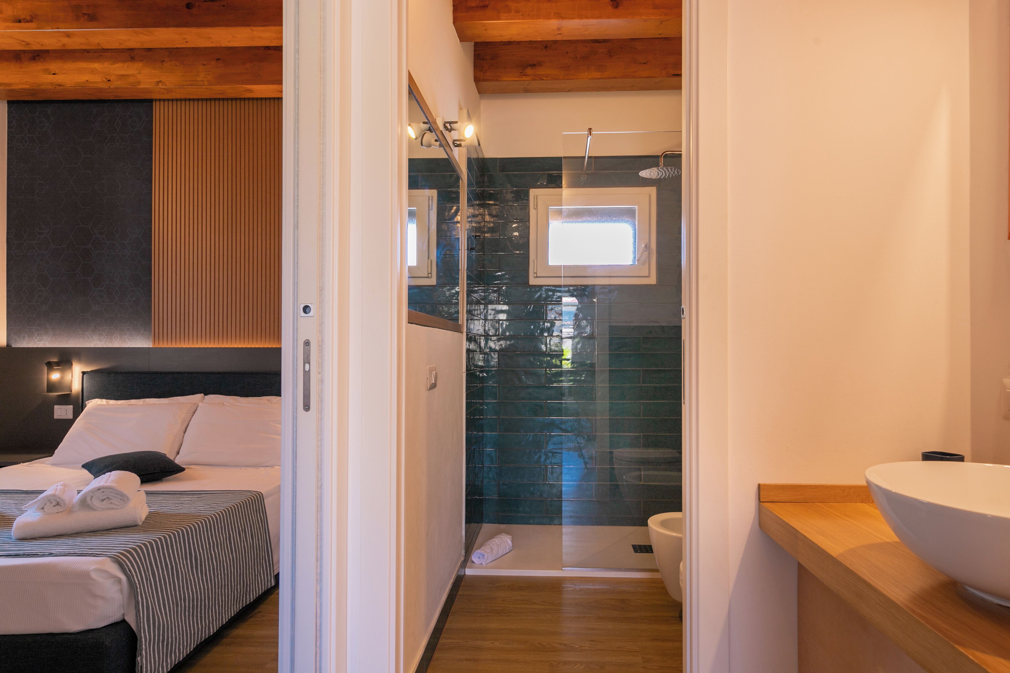 Una habitación de hotel moderna con una cama, cabecero de madera, un espejo grande, cabina de ducha con mampara de cristal y tocador de madera con lavamanos.