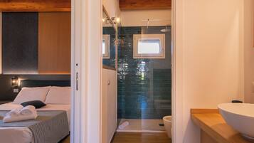 Una habitación de hotel moderna con una cama, cabecero de madera, un espejo grande, cabina de ducha con mampara de cristal y tocador de madera con lavamanos.