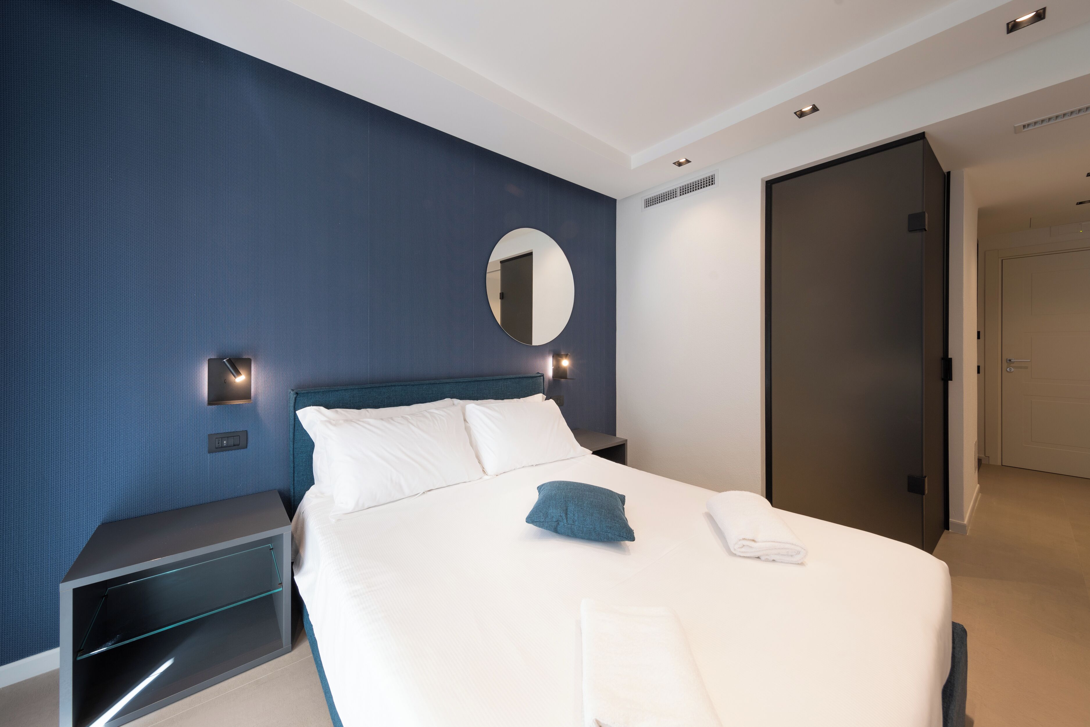 A modern hotel room with a large bed, a bedside table, a nightstand, a round mirror, and a door.