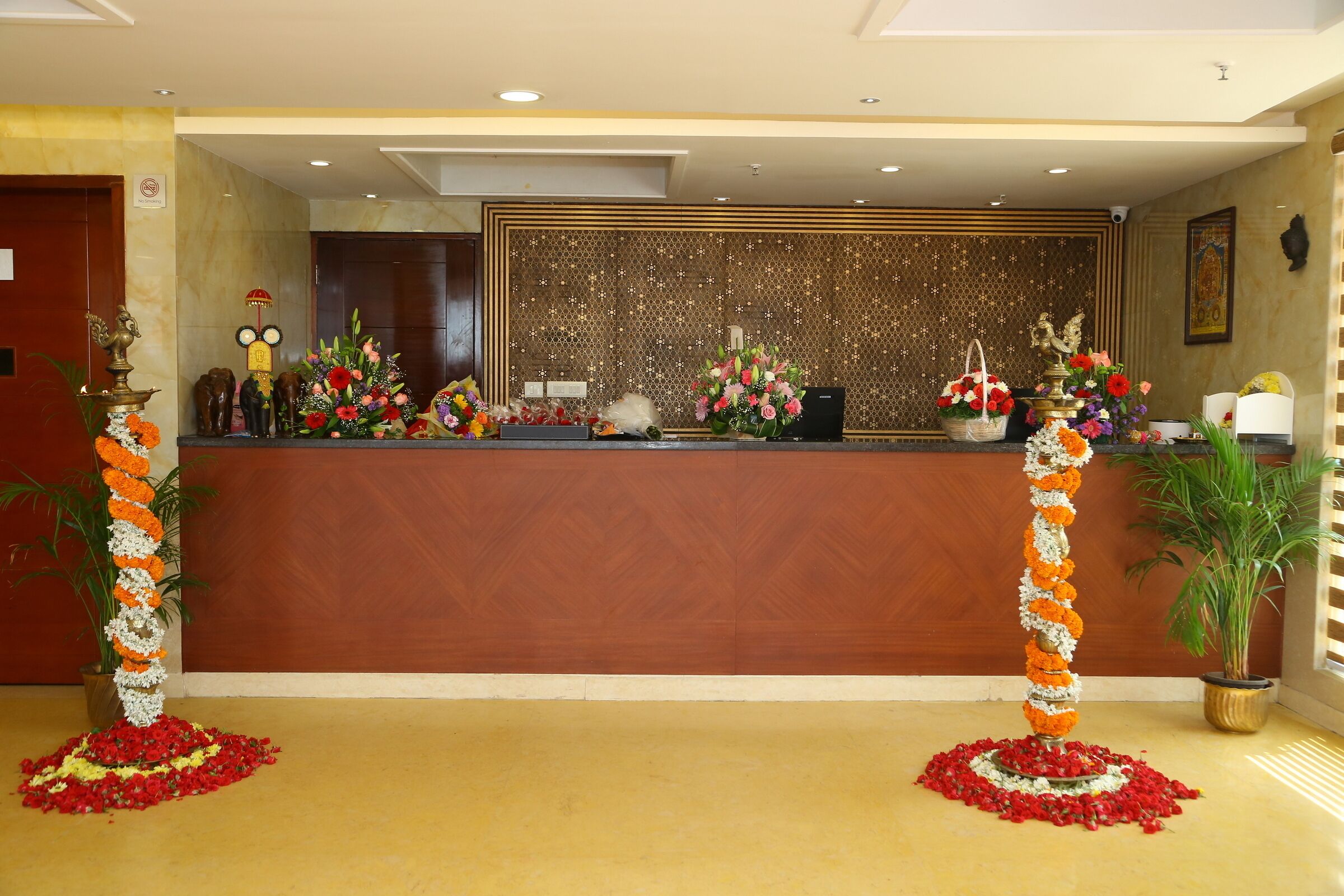 Un área de recepción con un mostrador de madera, una pared decorativa con un diseño geométrico y dos arreglos florales con base circular.