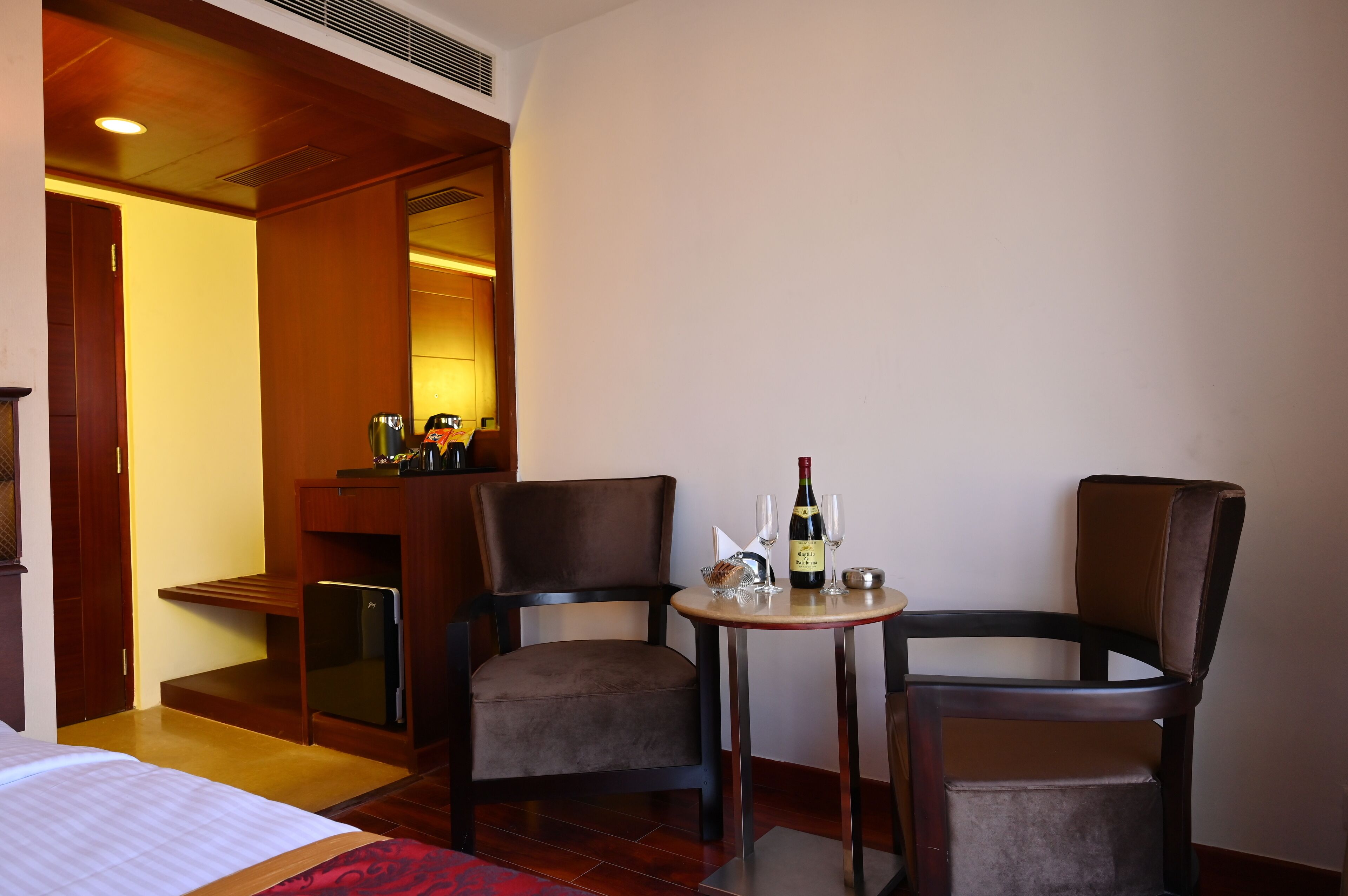 A hotel room with two chairs, a small table with a bottle and glasses, and a minibar.