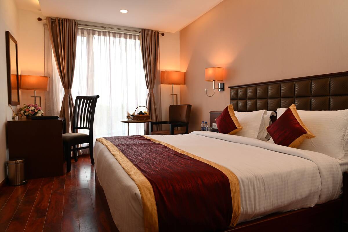 Habitación de hotel con una cama grande, dos mesitas de noche, un escritorio, una silla y una pequeña mesa con un jarrón de flores.