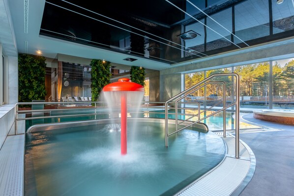 An indoor swimming pool with a water jet feature, surrounded by a modern architectural design with glass walls and a view of trees outside.