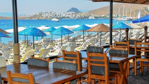Un restaurante frente a la playa con mesas y sillas de madera, sombrillas azules y vistas al mar y colinas al distante.