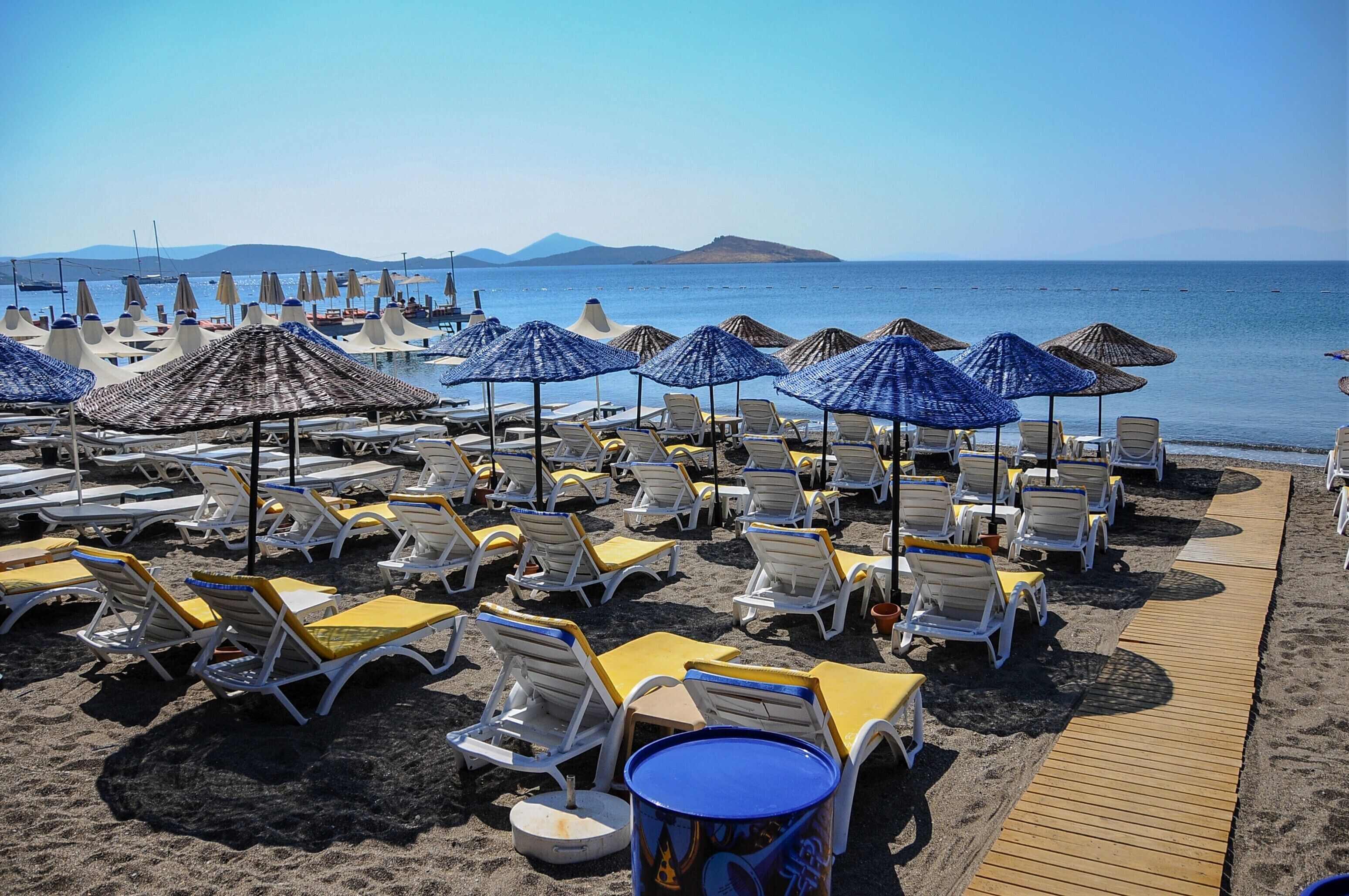 A beachside resort with rows of sun loungers and umbrellas, a wooden walkway, and a view of the sea and distant hills.