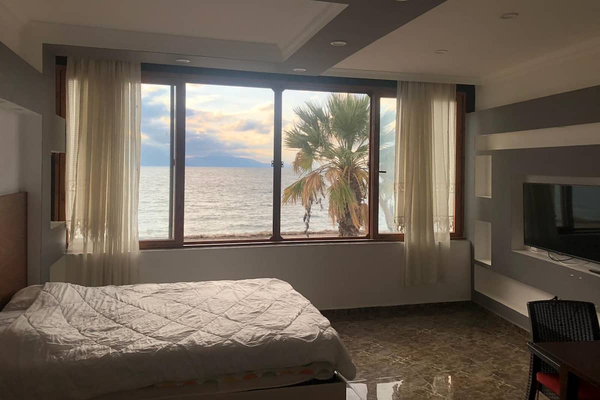 A bedroom with a large window overlooking the sea, a bed, a TV, and a desk.