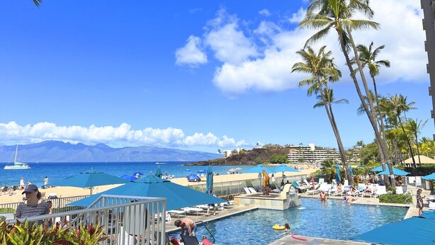 Un espace piscine d’un complexe hôtelier offrant une vue sur l’océan et les montagnes, entouré de palmiers et de parasols de plage.