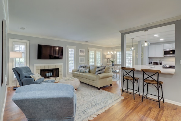 A modern living room with a fireplace, a sofa, and a TV. There is a kitchen area with bar stools and a dining table.