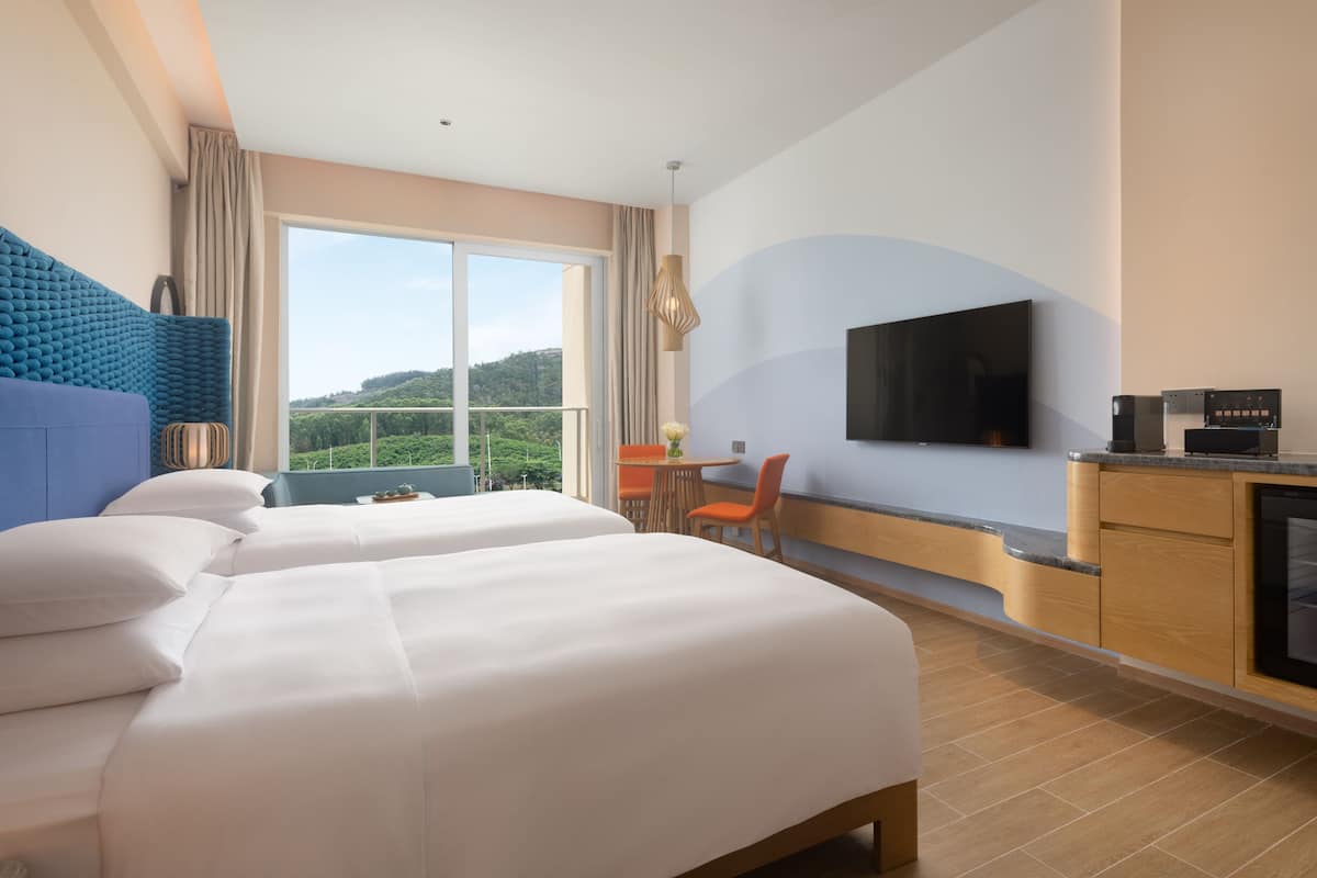 A hotel room with two beds, a TV, a small table with a chair, and a view of the outdoors.