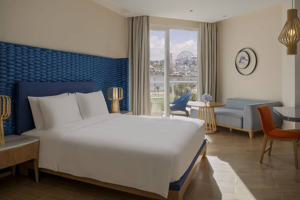 A modern hotel room with a large bed, a seating area with a table and chairs, and a view of a Ferris wheel through the window.