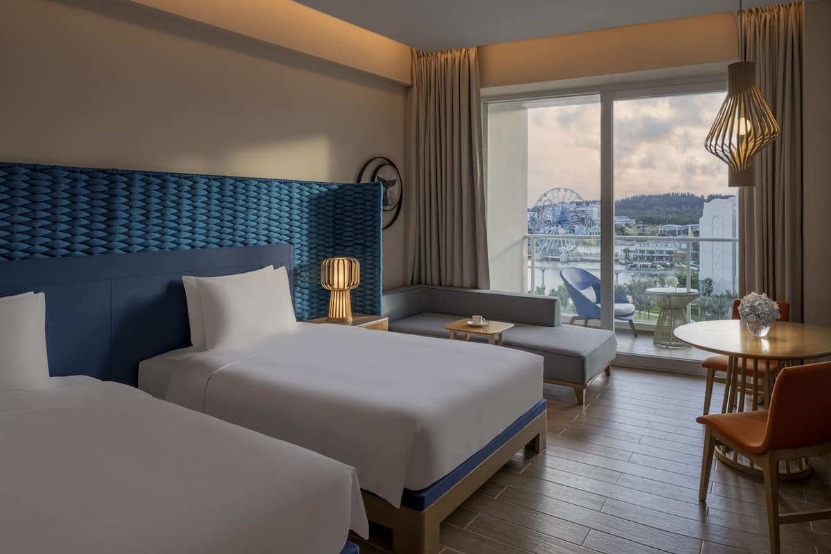 A hotel room with two beds, a sofa, a dining table, and a view of a cityscape and amusement park.
