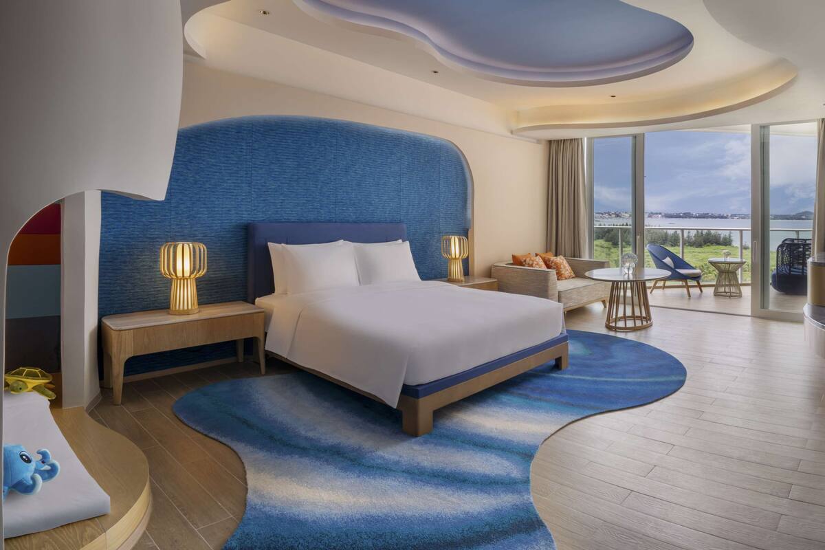 A modern hotel room with a large bed, a curved blue rug, a wooden bench, and a view of the outdoors.